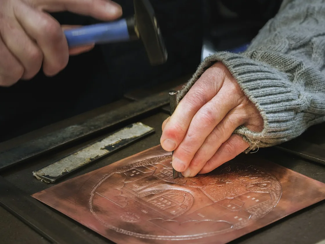 Manos trabajando con martillo y cincel trazador sobre una chapa de cobre con un dibujo grabado en la superficie